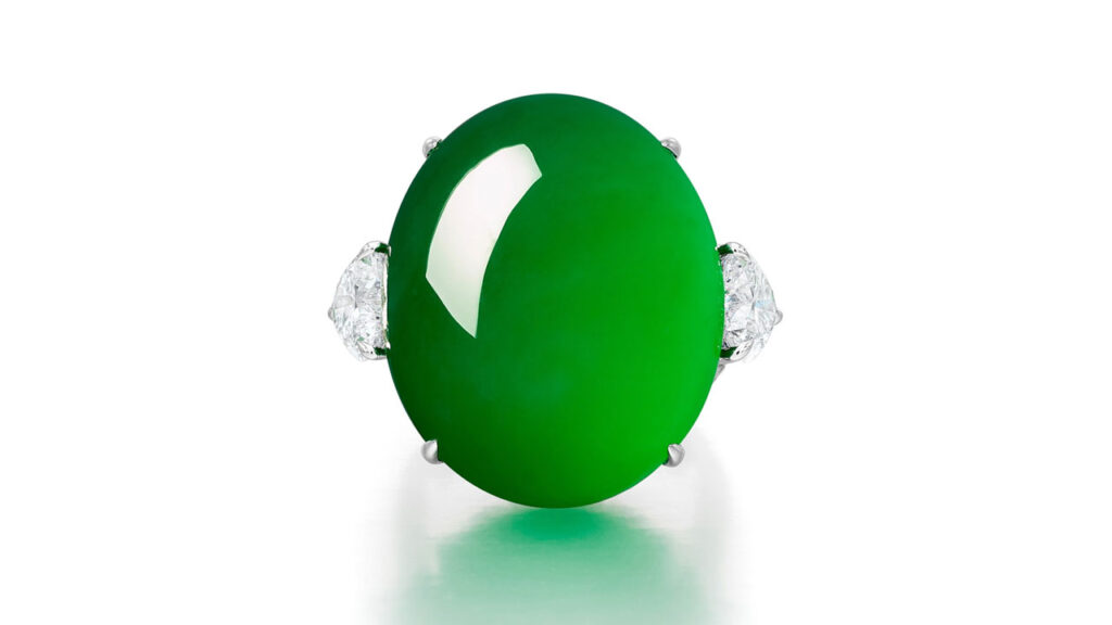 An oval jadeite cabochon ring of bright-green color, accented by two heart-shaped and brilliant-cut diamonds at Sotheby's image