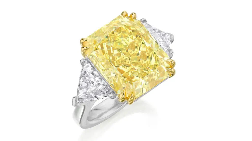 A ring bearing a cut-cornered rectangular modified brilliant-cut, 14.71-carat, fancy-intense-yellow, VS1-clarity diamond between two triangular-cut diamonds at Phillips image