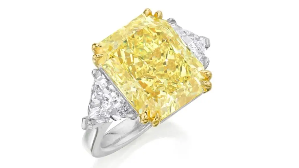 A ring centering a cut-cornered rectangular modified brilliant-cut, 14.71-carat, fancy-intense-yellow, VS1-clarity diamond, between two triangular-cut stones at Phillips image