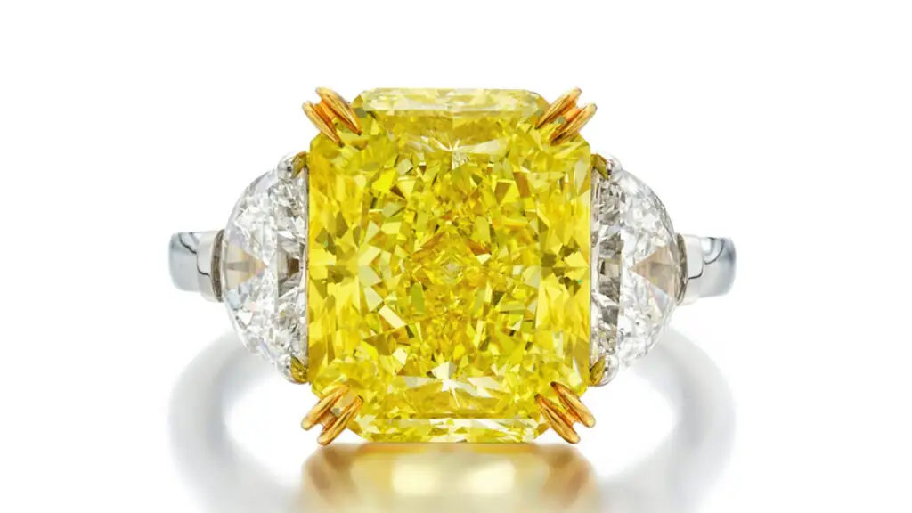 A ring features a cut-cornered rectangular modified brilliant-cut, 6.82-carat, fancy-vivid-yellow diamond between a pair of half-moon diamonds by Sotheby's image